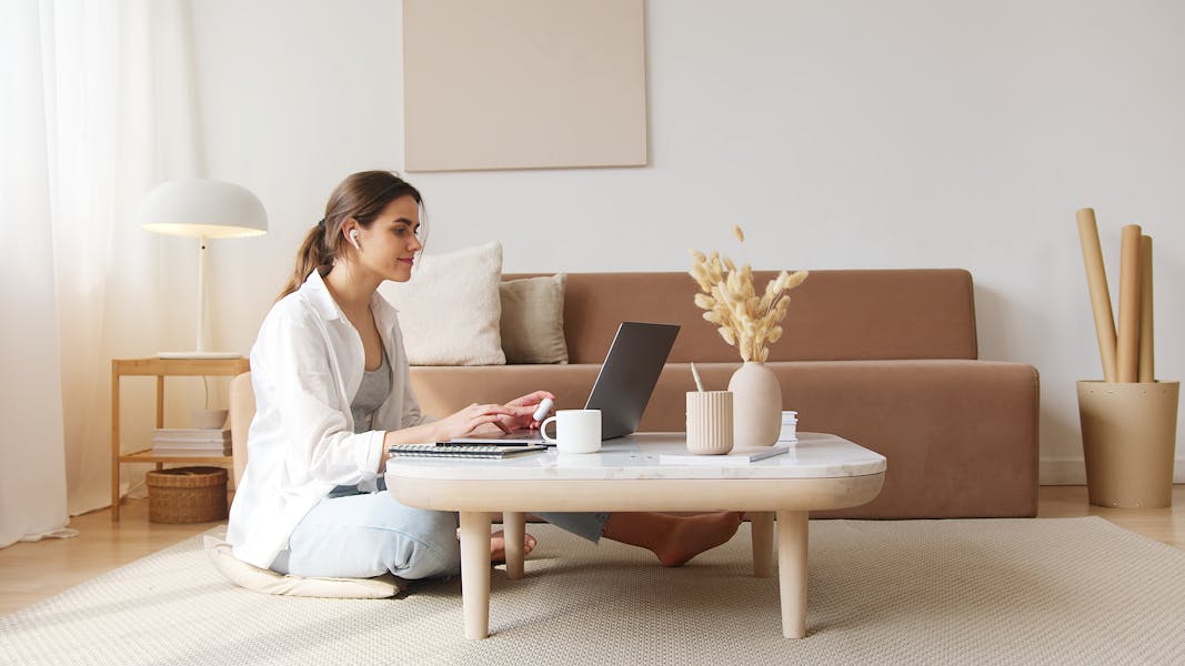 A woman uses a laptop at a coffee table in a cozy living room, embodying a relaxed work-from-home lifestyle.